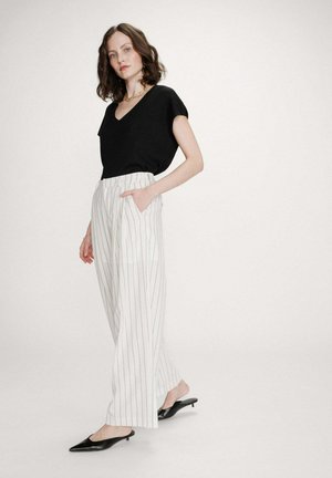 Femme aux cheveux bruns mi-longs portant un haut noir à manches courtes et col en V, un pantalon blanc à larges jambes rayé, des talons noirs pointus et un collier en or.