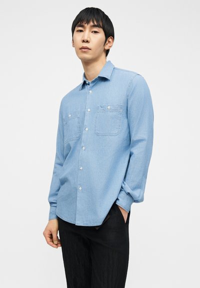 Νεαρός άνδρας με μαύρα μαλλιά, που φοράει ανοιχτό μπλε πουκάμισο με μακριά μανίκια και μαύρο παντελόνι, στέκεται με το ένα χέρι στην τσέπη.
