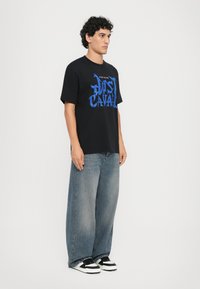 Fekete pamut póló nagy, kék "Just Cavalli" grafikával, széles, fakó kék farmernadrággal és fekete-fehér sneaker cipővel párosítva.