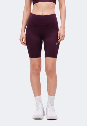Frau trägt dunkelviolette hoch taillierte Radlerhosen, weiße Socken und weiße Sportschuhe und steht vor einem schlichten Hintergrund.