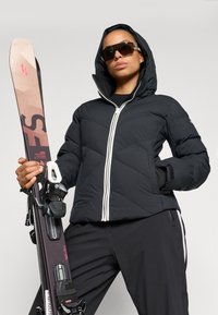 Sort pufferjakke med hætte, der har en hvid lynlås og chevron-syninger. Modellen holder et par ski med sorte bindinger. Solbriller bæres.