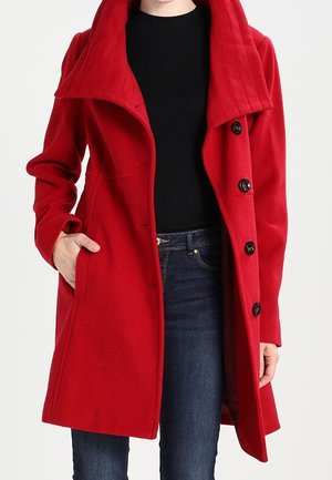 Woman wearing a bright red double-breasted coat with large collar, black top, and blue jeans, with one hand in coat pocket.