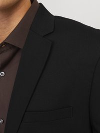 Black blazer with sharp lapels, tailored fit, and a pocket. Worn over a dark brown button-up shirt with a smooth texture and visible buttons.