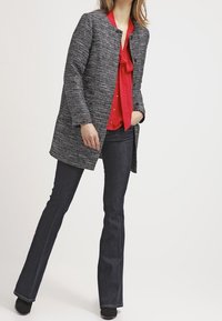 Grå texturerad kappa med rund halsringning, lagrad över en röd blus med rosetthals, kombinerad med mörka utsvängda jeans och svarta ankelstövlar.