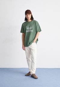 Zielona, oversize bawełniana koszulka z białym logo Adidas, w połączeniu z kremowymi spodniami i brązowymi zamszowymi butami, stojąca na niebieskim dywaniku.