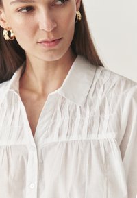 Blusa bianca a maniche lunghe con colletto. Presenta dettagli a righe sottili sul petto e un tessuto leggero e morbido.
