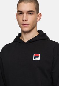 Musta huppari, jossa on etupuolella valkoinen, punainen ja sininen logo-patch, joka näyttää teksturoidulta, sekä kengurupocket. Kangas vaikuttaa pehmeältä.