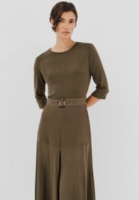 Oliwkowa sukienka z gładkiego materiału, okrągłym dekoltem, rękawami o długości trzech kwart, i paskiem w talii z prostokątną klamrą.