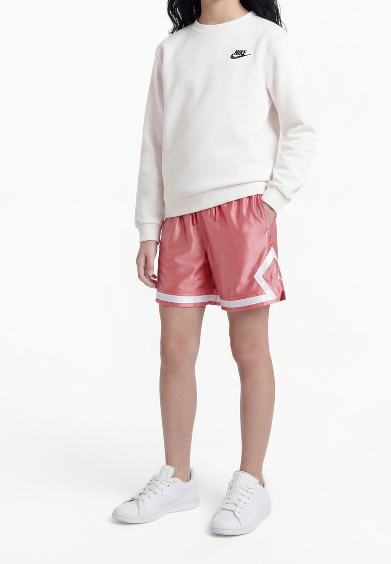 Jeune personne portant un sweat-shirt blanc Nike, un short de sport rouge avec des accents blancs et des baskets blanches, debout les mains dans les poches.