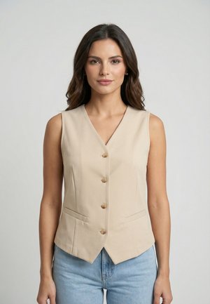 Donna con capelli scuri e ondulati che indossa un gilet beige abbottonato e jeans azzurri, in piedi davanti a uno sfondo grigio chiaro uniforme.