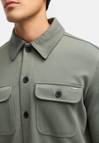 Grøn, struktureret jakke med knapkrave, to forlommer og sorte knapper. Har et metal-logo som accent på lommen.