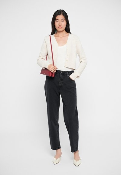 Kremfarget cardigan med knapper, brukt over en hvit topp, kombinert med mørke jeans og hvite spisse hæler. En liten rød crossover-veske kompletterer antrekket.