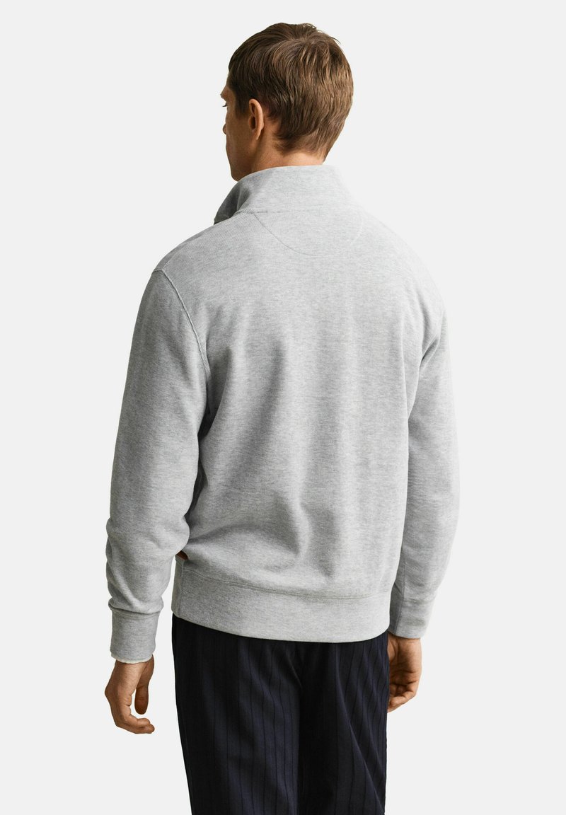 Mann von hinten, trägt einen hellgrauen Sweatshirt mit Rippenkragen und dunkel gestreifte Anzughose vor weißem Hintergrund.