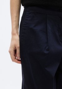 Marineblauwe broek met een gladde textuur, voorzien van een geplooide voorkant en een elastische tailleband. Hand geplaatst bij de zijnnaad.