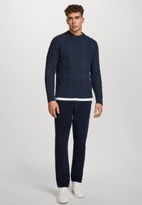 Granatowy sweter z dzianiny o teksturowanym wzorze w warkocze, okrągły dekolt i ściągacze przy rękawach, zestawiony z ciemnymi spodniami i białymi sneakerami.