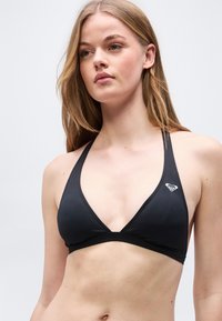 Νεαρή γυναίκα με μακριά ξανθά μαλλιά που φοράει μαύρο μαγιό τοπ halter με ένα μικρό λευκό λογότυπο στην αριστερή πλευρά.