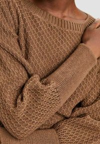 Pull tricoté marron texturé avec poignets et encolure côtelés porté par une personne avec la main posée sur l'épaule.