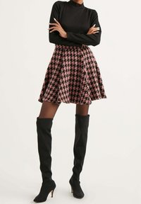 Schwarzes geripptes Langarmoberteil kombiniert mit einem flieder- und schwarzen Hahnentrittmuster-Rock mit weitem Schnitt. Das Model trägt kniehohe schwarze Stiletto-Stiefel.