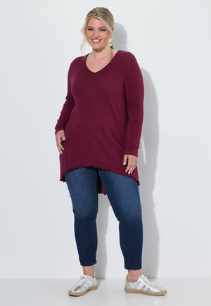 Glunderende vrouw met een lange bordeauxrode trui, donkerblauwe spijkerbroek en zilveren sneakers, staand tegen een effen lichte achtergrond.