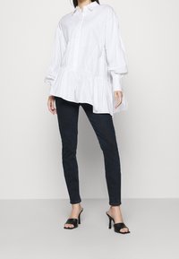 Vit bomullsskjorta med volangkant, knäppningsdesign och långa puffärmar, kombinerad med mörkblå slim fit-byxor och svarta klackskor.