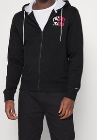Felpa nera con zip in cotone, con cappuccio foderato in grigio, caratterizzata da un logo rosso "TOMMY JEANS" sul petto e due tasche frontali.
