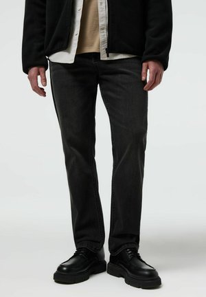 Mężczyzna w czarnych dżinsach, czarnych masywnych skórzanych butach, beżowej koszuli pod białą koszulą na guziki oraz czarnej kurtce na suwak, stojący na białym tle.