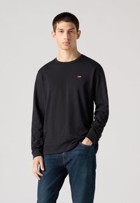 Černé bavlněné tričko s dlouhým rukávem a kulatým výstřihem, s malým červeným logem na hrudi. Kombinováno s modrými džínami.