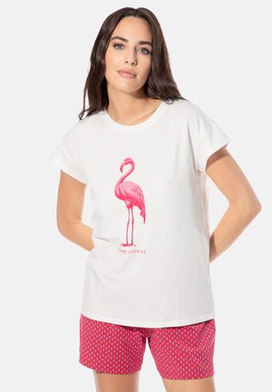 Frau trägt ein weißes T-Shirt mit pinkem Flamingo-Graphic und dem Text "Short Stories", kombiniert mit roten Shorts mit Muster, steht mit den Händen hinter dem Rücken.