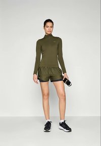 Langermet, olivengrønn treningsgenser med kvart glidelås, kombinert med svarte treningsshorts. Modellen holder en svart vannflaske og har på seg svarte joggesko.