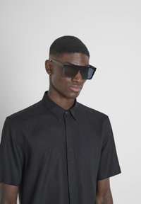 Gafas de sol rectangulares negras con lentes tintados, montura gruesa y acabado mate, llevadas con una camisa negra de manga corta y botones.