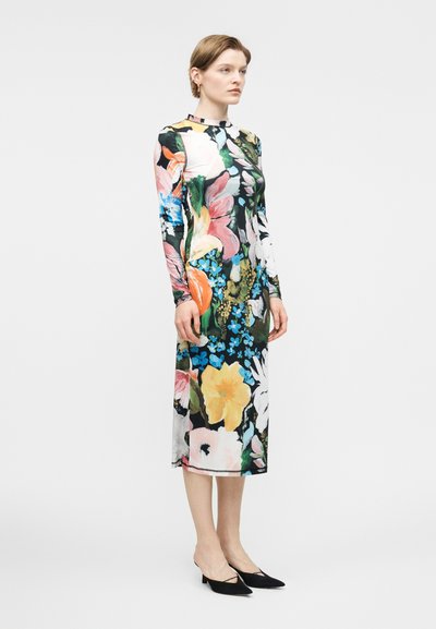 Γυναίκα που φοράει μακρυμάνικο, μίντι φόρεμα με πολύχρωμα λουλούδια σε έντονες αποχρώσεις μπλε, κίτρινου, ροζ και λευκού, συνδυασμένο με μαύρες μυτερές γόβες.