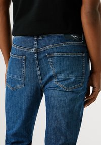 Modré denimové džíny s úzkým střihem, mají zadní kapsy a tmavou koženou nášivku na pase. Bledé švy podrobně zpracované.