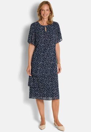 Lächelnde Frau, die ein marineblaues, knielanges Kleid mit weißen Punkten und kurzen Rüschenärmeln trägt, kombiniert mit beigen, niedrigen Absatzschuhen.