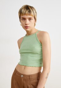 Ribbet grønn halterneck crop topp laget av strikket stoff, med en bølget kant og en liten metallisk detalj, kombinert med brune høyt midje bukser.