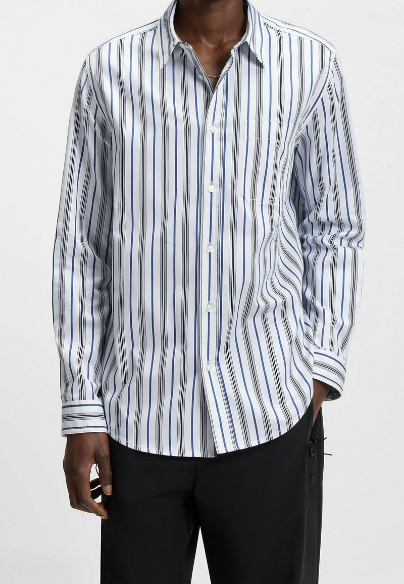 Homme portant une chemise blanche à boutons avec des rayures verticales noires et bleues, associée à un pantalon noir, debout devant un fond uni.