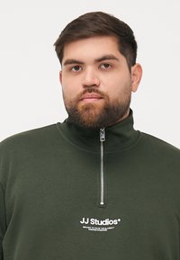 Green zip-up sweatshirt made from soft fabric. Features a high collar and a front zipper. Displays white logo text on the chest.