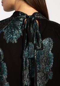 Zwarte blouse met een metallic teal bloemenpatroon, voorzien van een hoge hals en een striksluiting aan de achterkant, die een textuurdesign heeft.