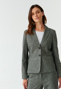 Šedý vlněný blazer s dokonalým střihem, klopovými límečky, dvěma černými knoflíky a bočními kapsami, doplněný bílým topem pod ním.