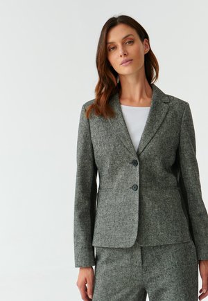 Grauer Wollblazer mit taillierter Passform, Reverskragen, zwei schwarzen Knöpfen und seitlichen Taschen, kombiniert mit einem weißen Top darunter.