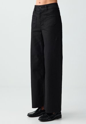 Czarne spodnie z wysokim stanem i szerokimi nogawkami z przednimi kieszeniami, noszone z czarnymi mokasynami, pokazane na modelce od talii do stóp na jednolitym tle.