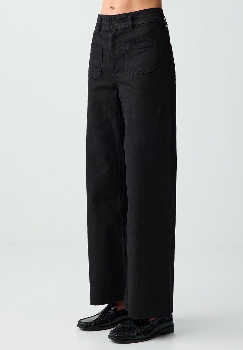Pantalon noir taille haute à jambes larges avec poches avant, porté avec des mocassins noirs, montré sur un mannequin de la taille aux pieds sur un fond uni.