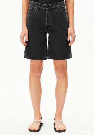 Personne portant un short en denim noir taille haute et des sandales blanches à brides, debout devant un fond blanc uni.