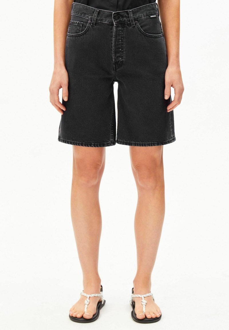 Person wearing black high-waist denim shorts and white strap sandals, standing against a plain white background.