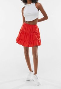 Kvinna iklädd en vit ärmlös crop top, röd lager-på-lager minikjol och vita sneakers stående mot en enfärgad vit bakgrund.