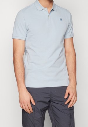 Mann trägt hellblaues Poloshirt mit kurzen Ärmeln und Kragen sowie dunkle Cargo-Hose, steht vor einfarbigem Hintergrund.