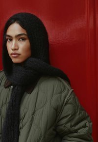 Gepolsterte olivgrüne Jacke mit dunklem gemustertem Schal, ergänzt durch einen glatten roten Hintergrund. Weiche Texturen, strukturierte Form.