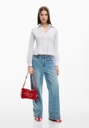 Frau mit einer taillierten weißen Bluse, weiten blauen Jeans, roten Schuhen und einer roten Handtasche mit geflochtenem Henkel vor weißem Hintergrund.