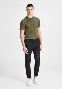 Olivgrünes Polohemd mit Kragen, kombiniert mit schwarzen Slim-Fit-Hosen und weißen Sneakers. Einfaches Design, kurze Ärmel, minimale Akzente.