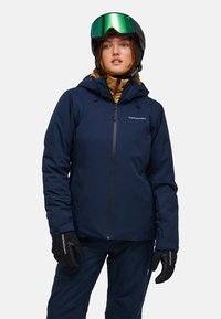 Frau trägt eine marineblaue Skijacke, passende Hose, schwarze Handschuhe, schwarzen Helm und grüne Skibrille und schaut leicht nach rechts.