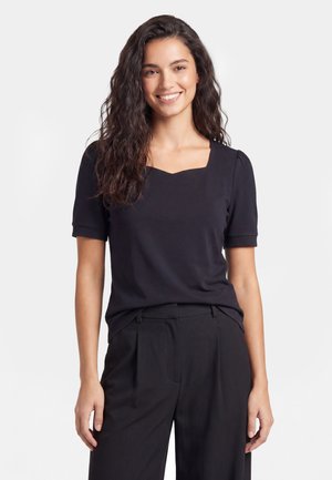 Femme aux longs cheveux ondulés sourit, portant un haut noir à manches courtes avec un col carré et un pantalon noir, debout devant un fond uni.